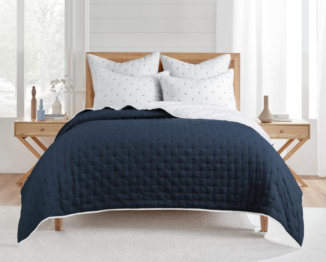 A dark blue coverlet set with white Swiss blue dots, displayed on a bed in a room with a wooden bed frame and white walls.